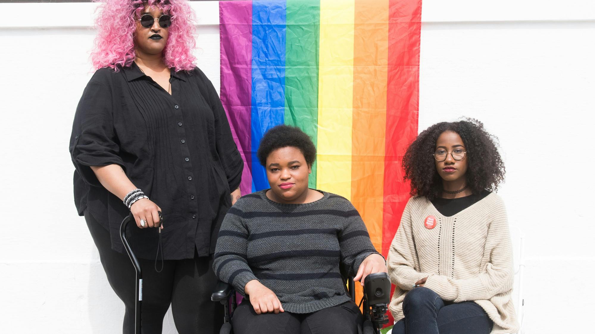 Three Black and disabled folx (a non-binary person holding a cane, a non-binary person  sitting in a power wheelchair, and a femme sitting in a chair) look seriously at the camera while a rainbow pride  flag drapes on the wall behind them. Photo attributed to Disabled and Here project.