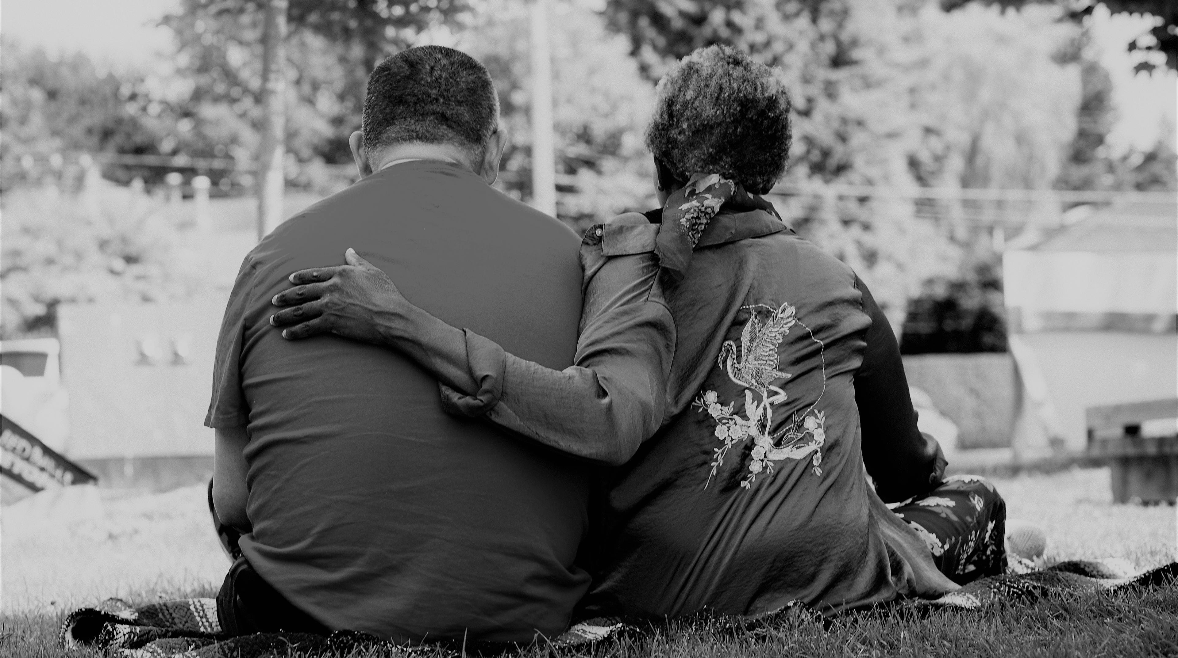 black and white picture of a man and woman sitting on a blanket in the grass with their backs to the camera and the woman's arm wrapped lovingly around the back of her partner.