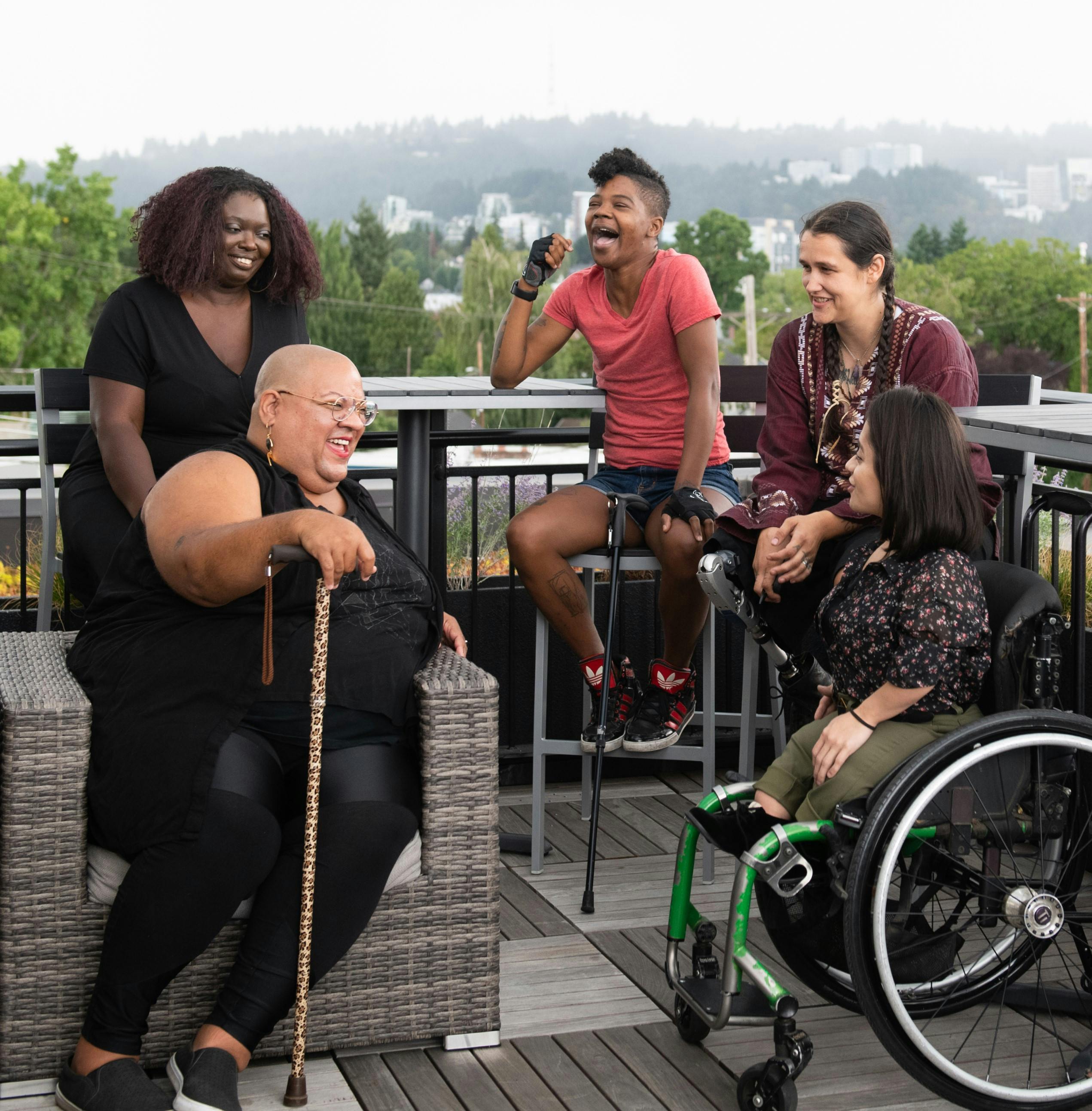 Five disabled people of color with canes, prosthetic legs, and a wheelchair sit on a rooftop deck,  laughing and sharing stories. Greenery and city high-rises are visible in the background. Photo attributed to Disabled and Here project.